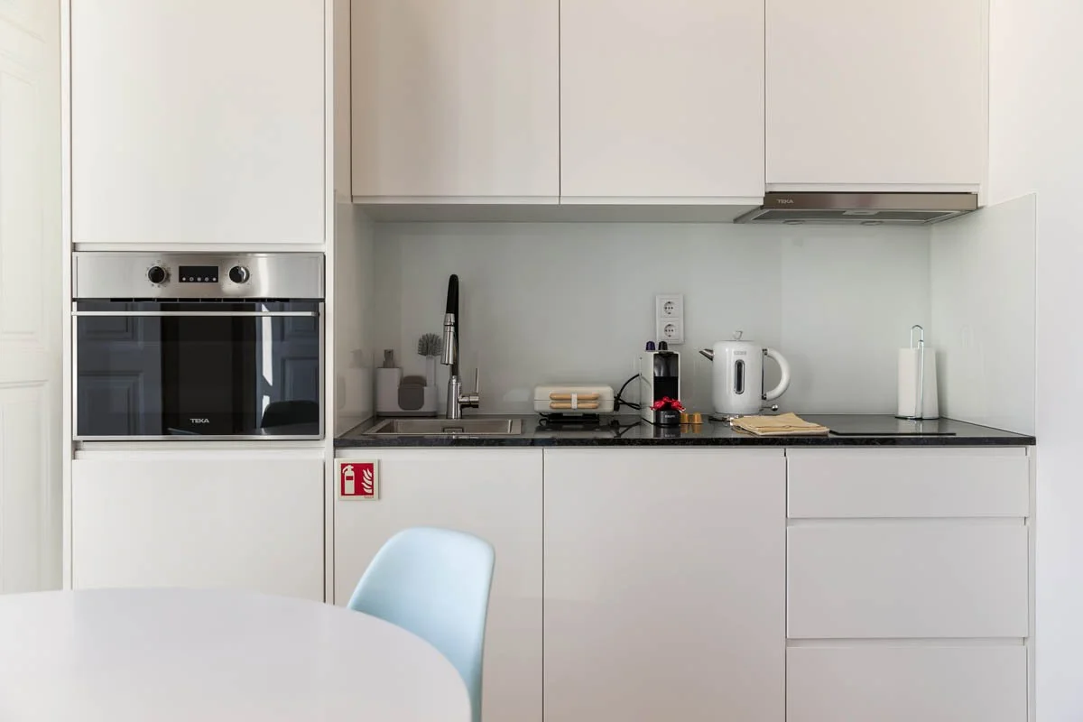 Cozinha com armários brancos, forno embutido, pia com torneira, eletrodomésticos como chaleira elétrica e máquina de café, e mesa com cadeira azul.