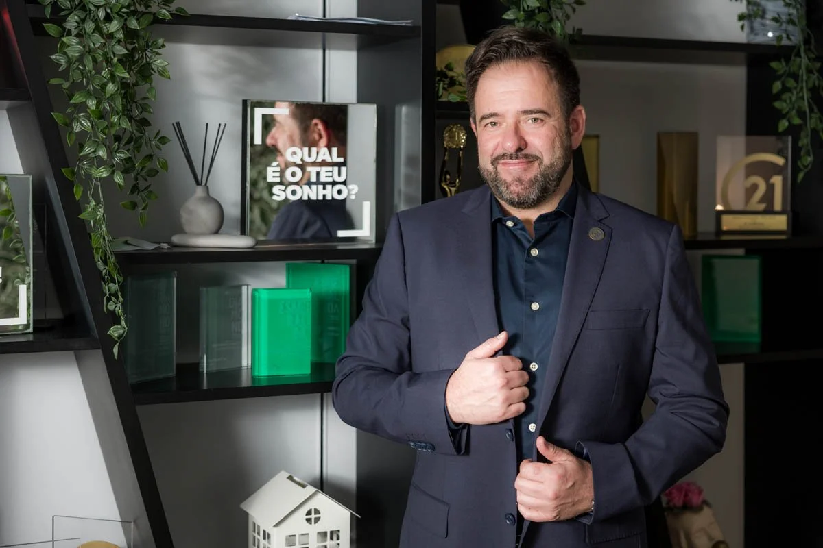 Homem de terno azul escuro posando em ambiente com decoração moderna, com livros, plantas e objetos decorativos ao fundo.
