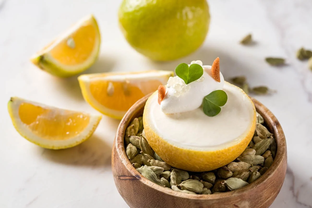 Laranja cortada ao meio com recheio de creme, decorada com folhas verdes e pedaços de bolo de amendoim. Em volta, fatias de limão e uma maçã verde ao fundo.