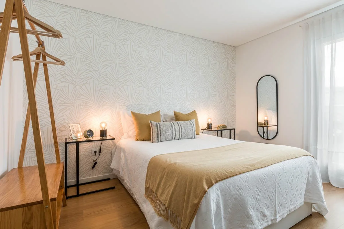 Quarto com parede de papel de parede branco com padrão de linhas douradas, cama com roupa branca, travesseiros bege e um travesseiro decorativo cinza com listras, duas mesinhas de cabeceira com luminárias acesas, espelho de corpo inteiro com moldura preta, janela com cortinas brancas.