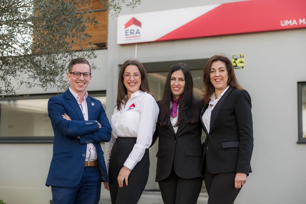 quatro pessoas casadas de negócios vestidas com roupas formais, duas mulheres de cabelos escuros e duas mulheres de cabelos castanhos, sorrindo e posando na frente de um prédio com sinal de ERA imobiliária.