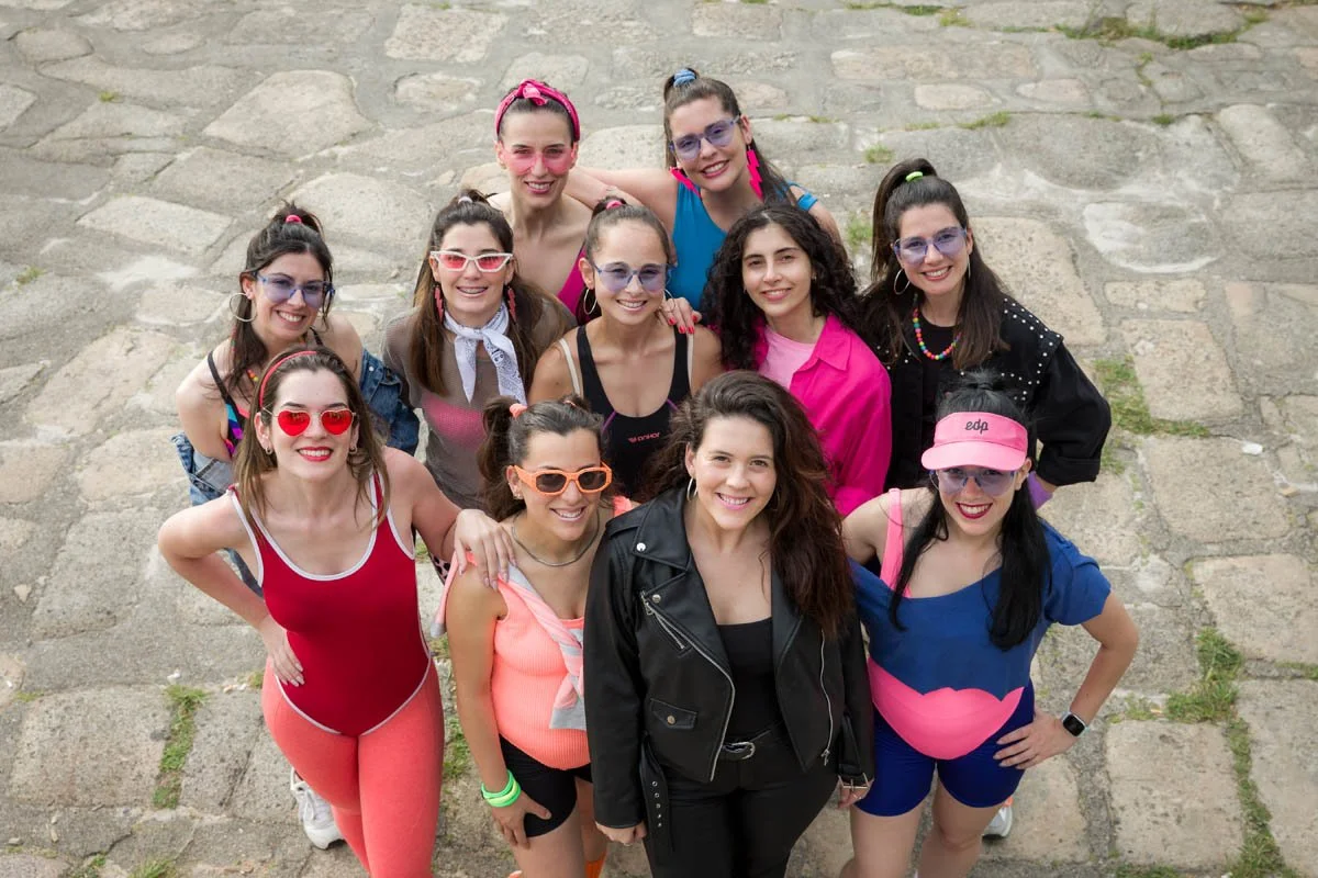 Grupo de jovens mulheres posando para a foto em uma rua de paralelepípedos, sorrindo e usando roupas coloridas e acessórios diversos.
