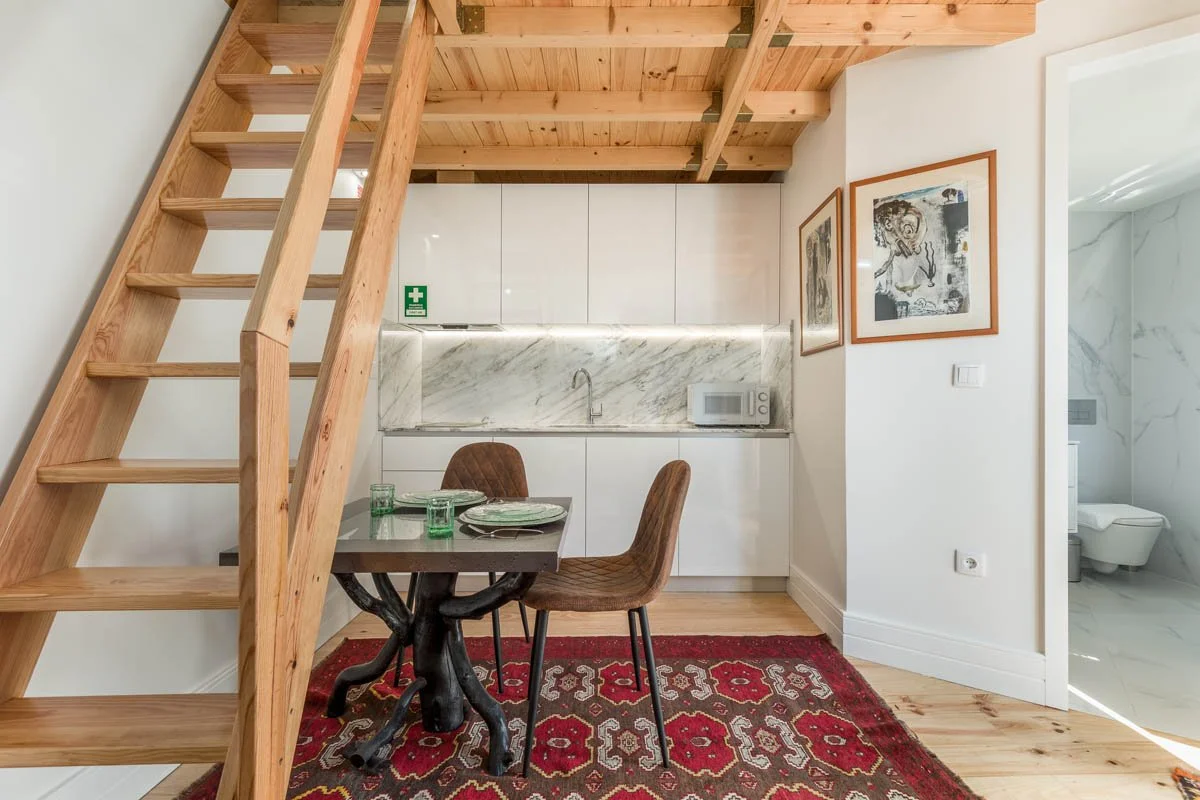 Interior de um apartamento com cozinha, área de jantar, escada de madeira, almofadas de veludo, tapete vermelho e uma porta levando ao banheiro com piso de mármore e vaso sanitário.