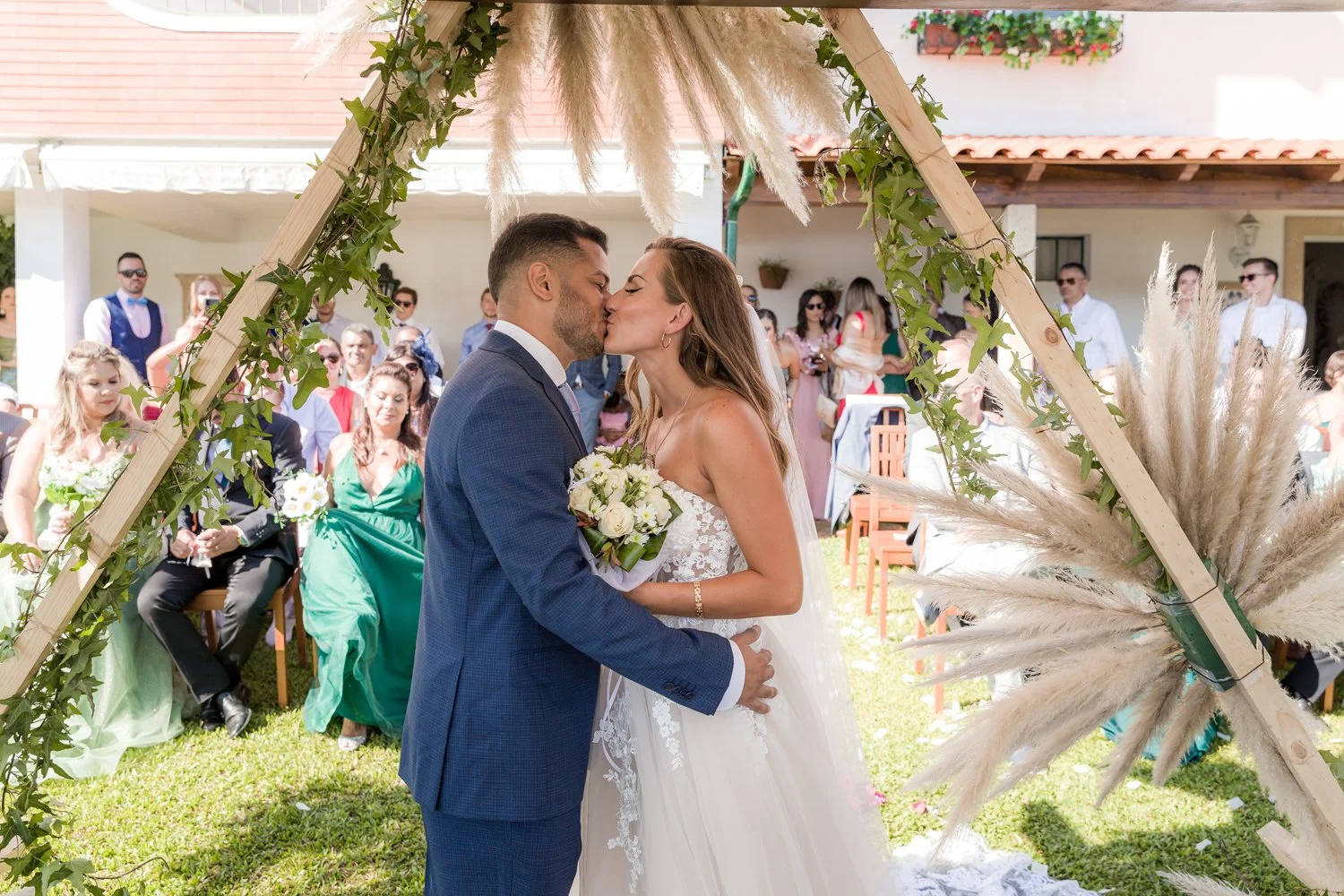 Casal de noivos se beijando na cerimônia de casamento ao ar livre, sob arco decorado com plantas e pampas, com convidados ao fundo.