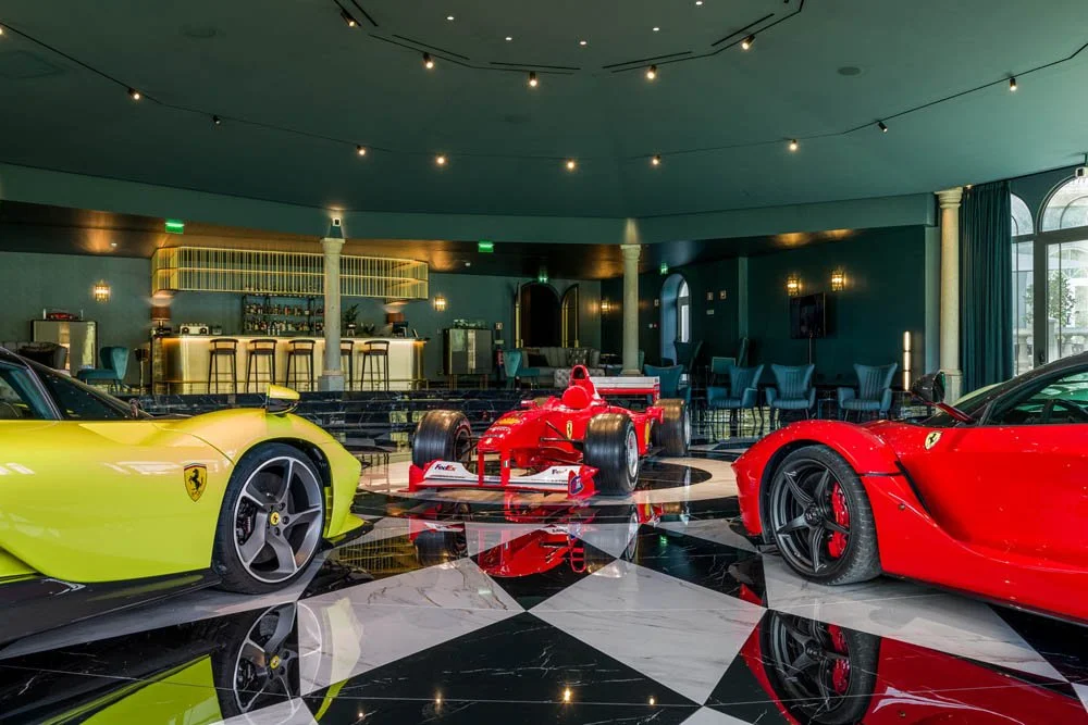 Interior de uma sala com carros de corrida Ferrari vermelhos e um carro de Fórmula 1 vermelho no centro, com piso preto e branco em padrão xadrez, paredes verdes, iluminação ambiente e área de bar ao fundo.