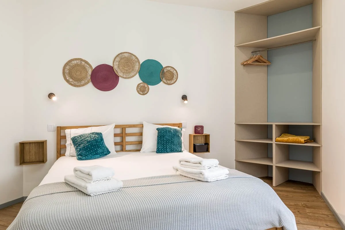 Quarto com cama de casal, cabeceira de madeira, duas almofadas verdes, roupa de cama branca, toalhas dobradas em cima da cama, parede decorada com círculos de palha e tecidos coloridos, parede branca, prateleira de canto com closet aberto com cabides