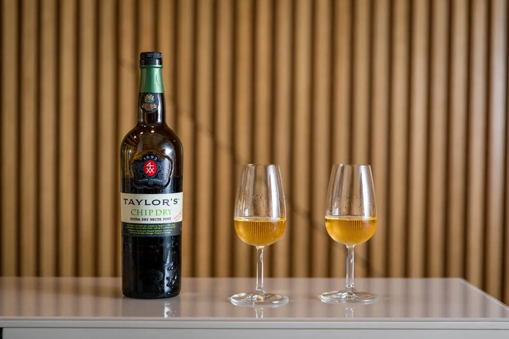 Garrafa de chá Taylor's Chip Dry ao lado de duas taças com bebidas amareladas, sobre uma mesa branca, com fundo de painéis de madeira vertical.