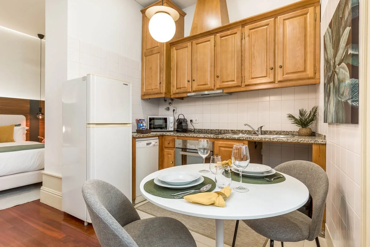 Cozinha com armários de madeira, micro-ondas, geladeira branca, mesa de jantar com pratos, copos, talheres e toalha de pano, em ambiente de decoração moderna.