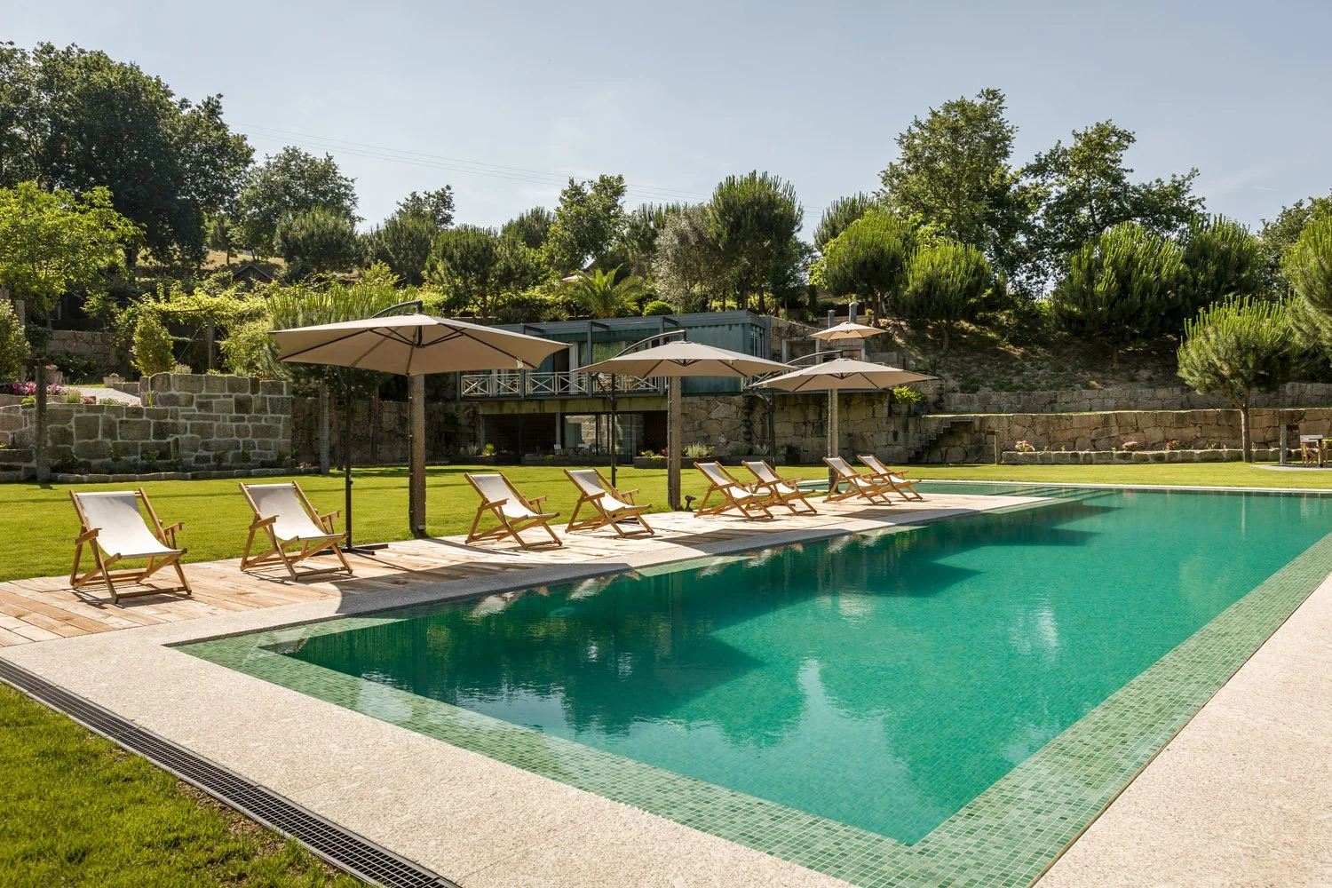 Piscina em um jardim com espreguiçadeiras e guarda-sóis de palha, cercada por vegetação e encosta com árvores.