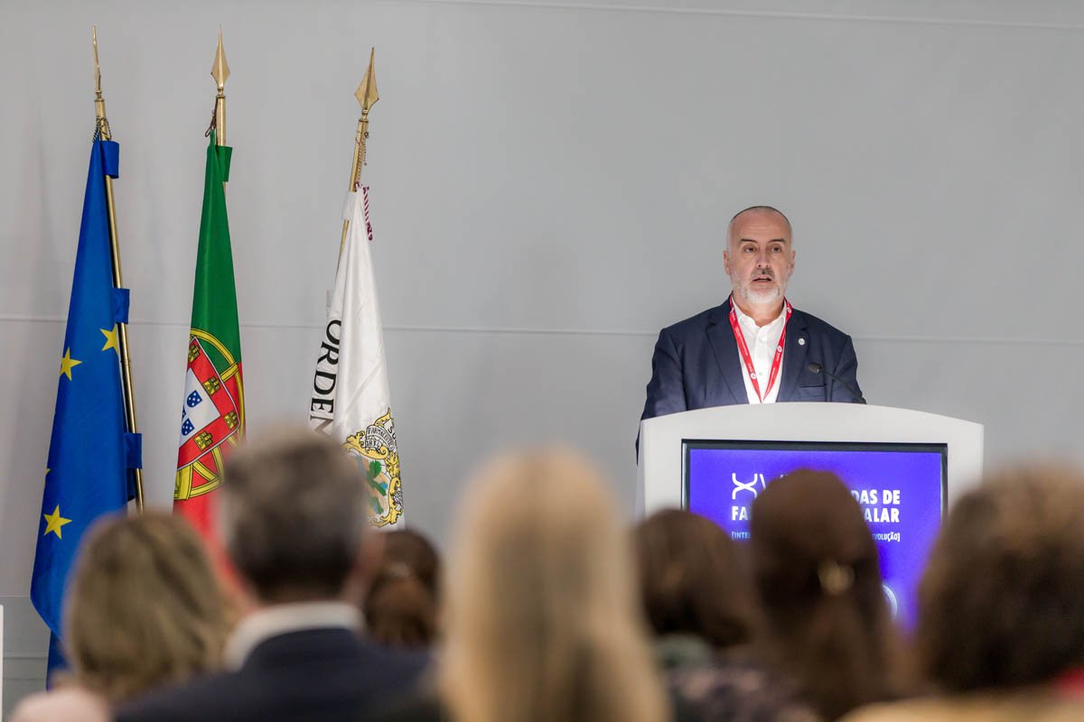 Homem falando em um palco com um púlpito, ao fundo há quatro bandeiras, incluindo a da União Europeia e Portugal, enquanto uma audiência assiste ao evento.