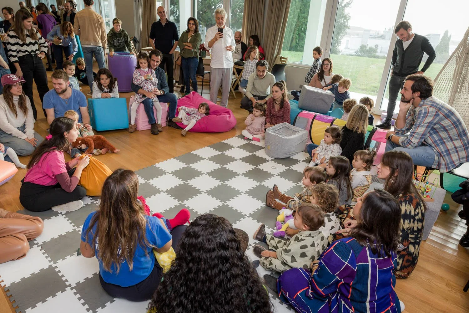 Pessoas, incluindo crianças, sentadas e assistindo a uma apresentação em um ambiente interno com janelas grandes e vista para o exterior, decorado com almofadas e tapete acolchoado.