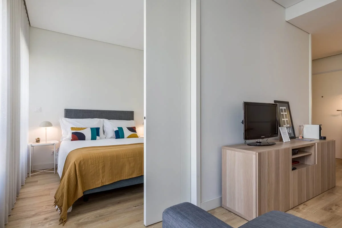 Quarto de hotel com cama de casal, cabeceira cinza, roupa de cama branca, almofadas coloridas, cobertor mostarda, abajur ao lado da cama, cortinas brancas, sala de estar com televisão, móvel de madeira, alguns quadros, objetos de decoração e aparelho