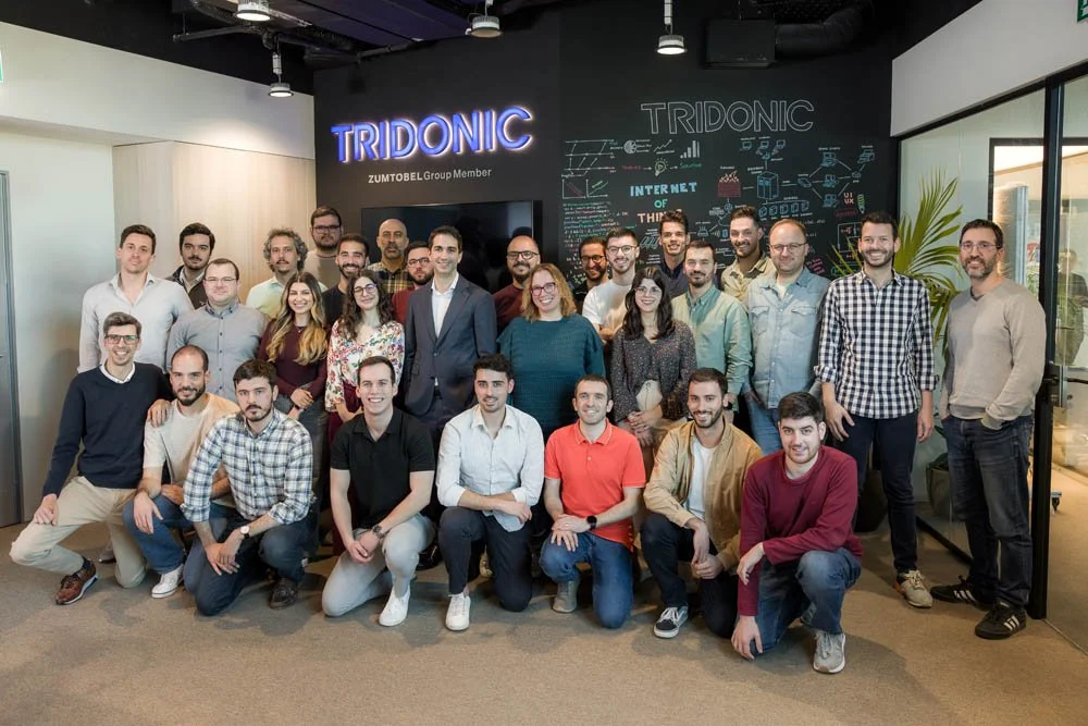 Grupo de pessoas posando para uma foto em ambiente de escritório, com um quadro negro ao fundo com desenhos de tecnologia e a palavra TRIDONIC em destaque
