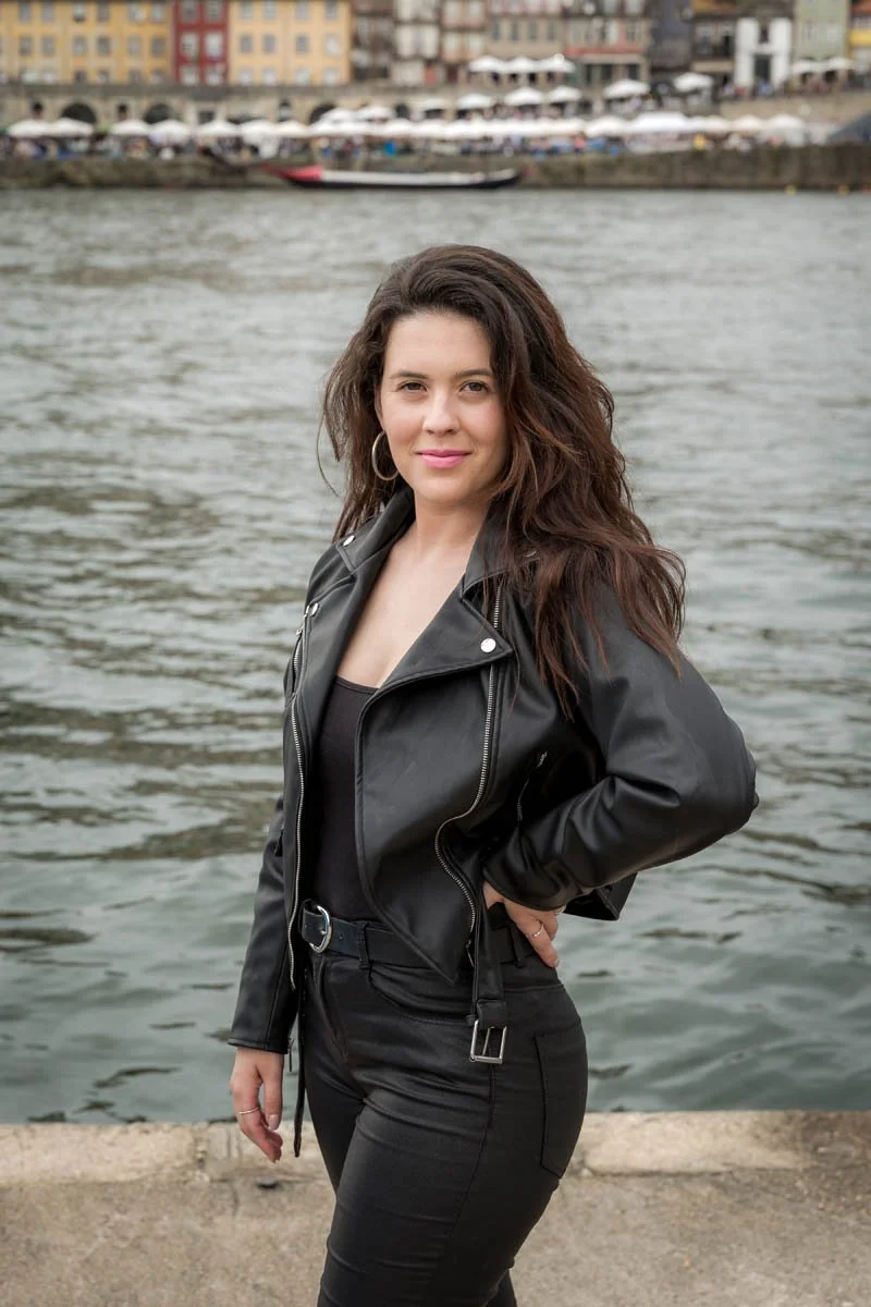 Mulher de cabelos longos e castanhos, usando jaqueta de couro preta e jeans pretos, posando na beira de um rio com prédios coloridos ao fundo.