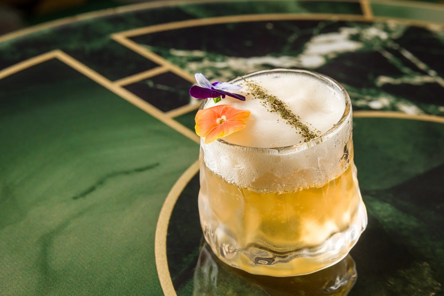 Copo de coquetel dourado com espuma na superfície, decorado com flores com cores roxa, rosa e laranja, colocado sobre uma mesa com padrão vegetal verde e detalhes dourados.