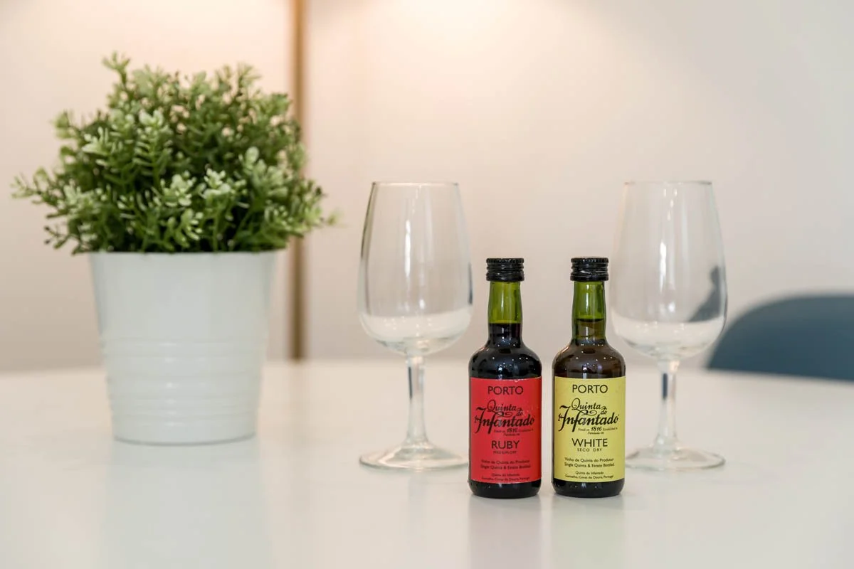 Duas garrafas de vinho do Porto, uma com rótulo vermelho e outra amarela, acompanhadas de duas taças de vinho vazias, ao lado de uma planta em vaso branco em uma mesa branca.