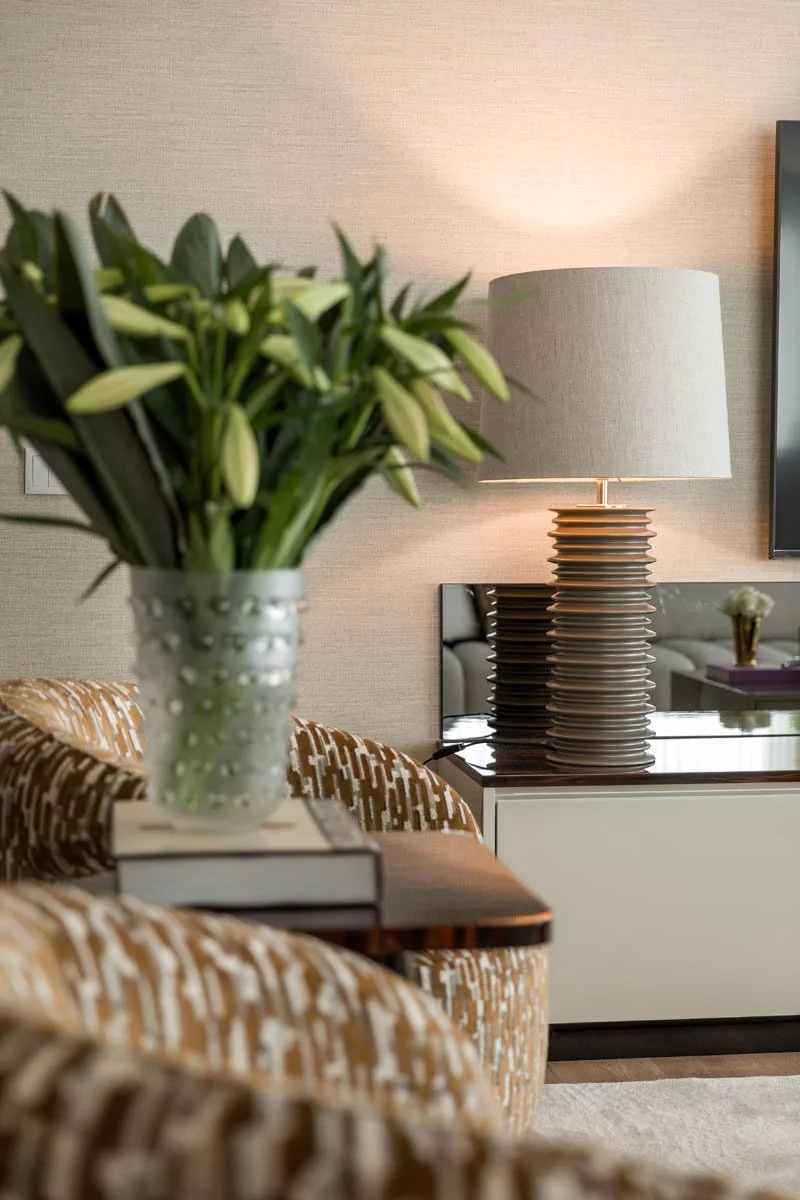 Arranjo de flores com lírios brancos em vaso decorativo em primeiro plano em um ambiente de sala com mesa de canto, mesinhas laterais, lampada de mesa e televisão.