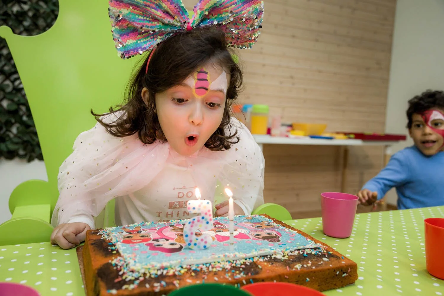 Menina soprando as velas do bolo de aniversário decorado com tema de festa infantil em uma mesa verde com ímãs coloridos, ao lado de outro menino com maquiagem facial, em um ambiente de celebração