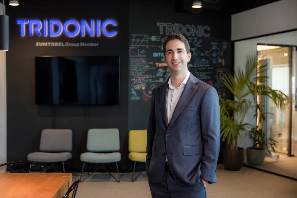 Homem de terno sorrindo em ambiente de escritório, com quadro negro e cadeira ao fundo, marca Tridonic na parede.