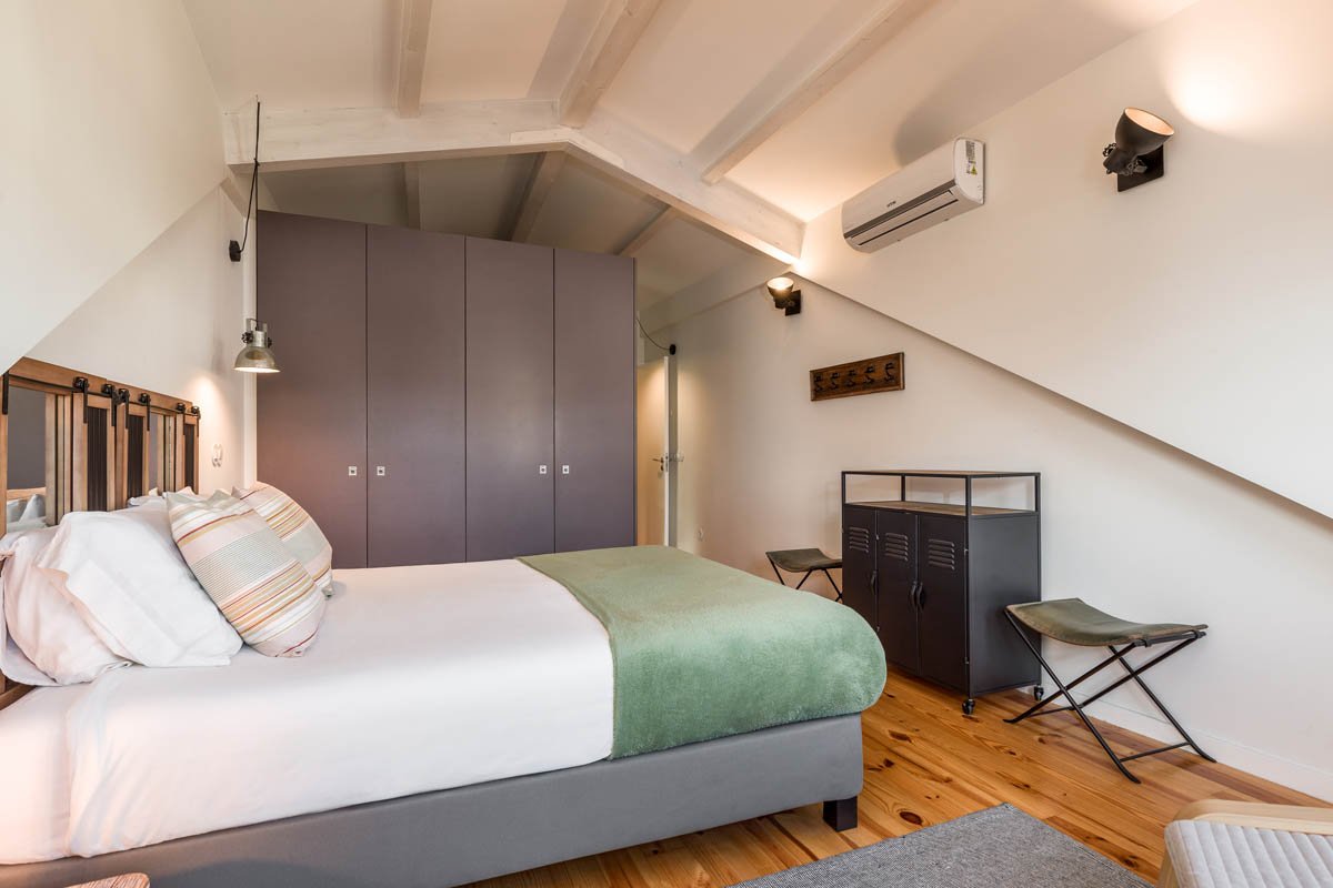 Quarto com cama de casal com roupa branca e travesseiros listrados, parede com closet de portas cinzas, piso de madeira, parede de teto inclinada com iluminação embutida e ar-condicionado, banco e móveis de metal na parede oposta.