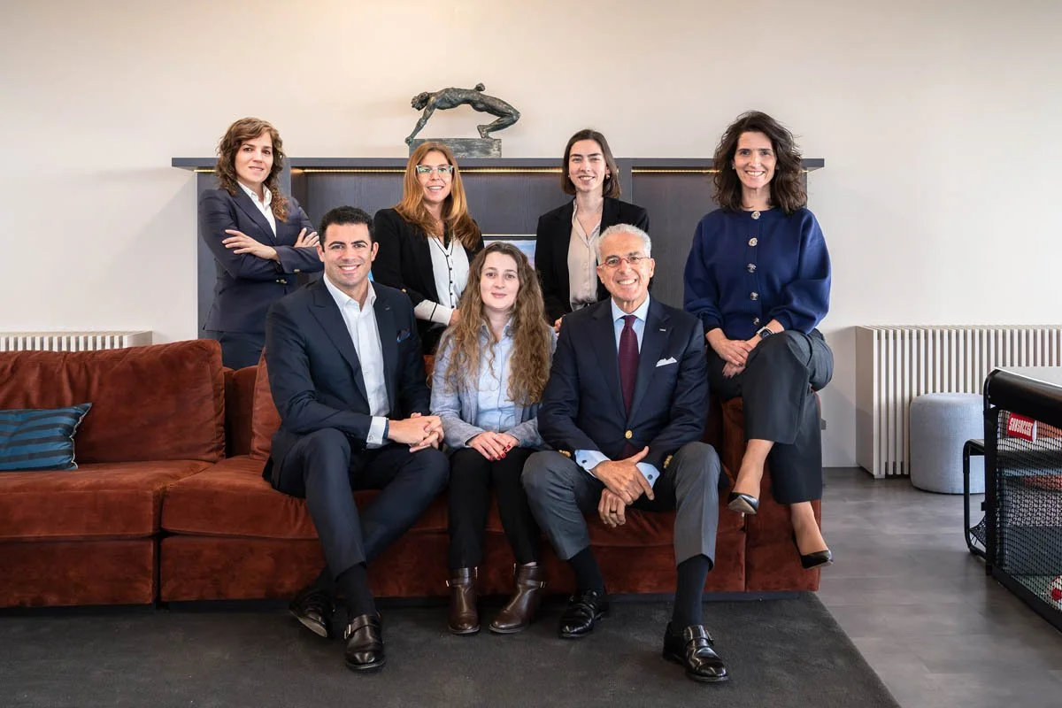 Grupo de sete pessoas, cinco mulheres e dois homens, posando sentados e de pé em um sofá de veludo marrom na sala de uma casa ou escritório moderno com decoração minimalista.