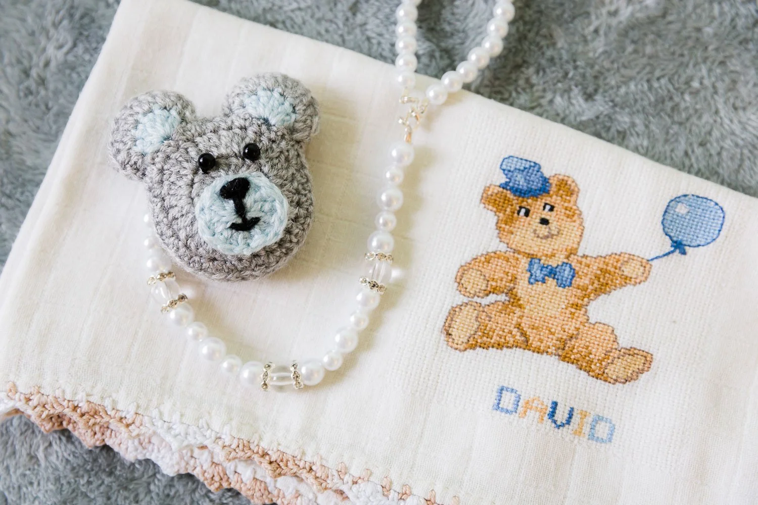 Urso de croche cinza com detalhes em azul, colar de pérolas, e uma toalha com desenho de urso de tricô segurando um balão, tudo sobre uma superfície de textura felpuda cinza.