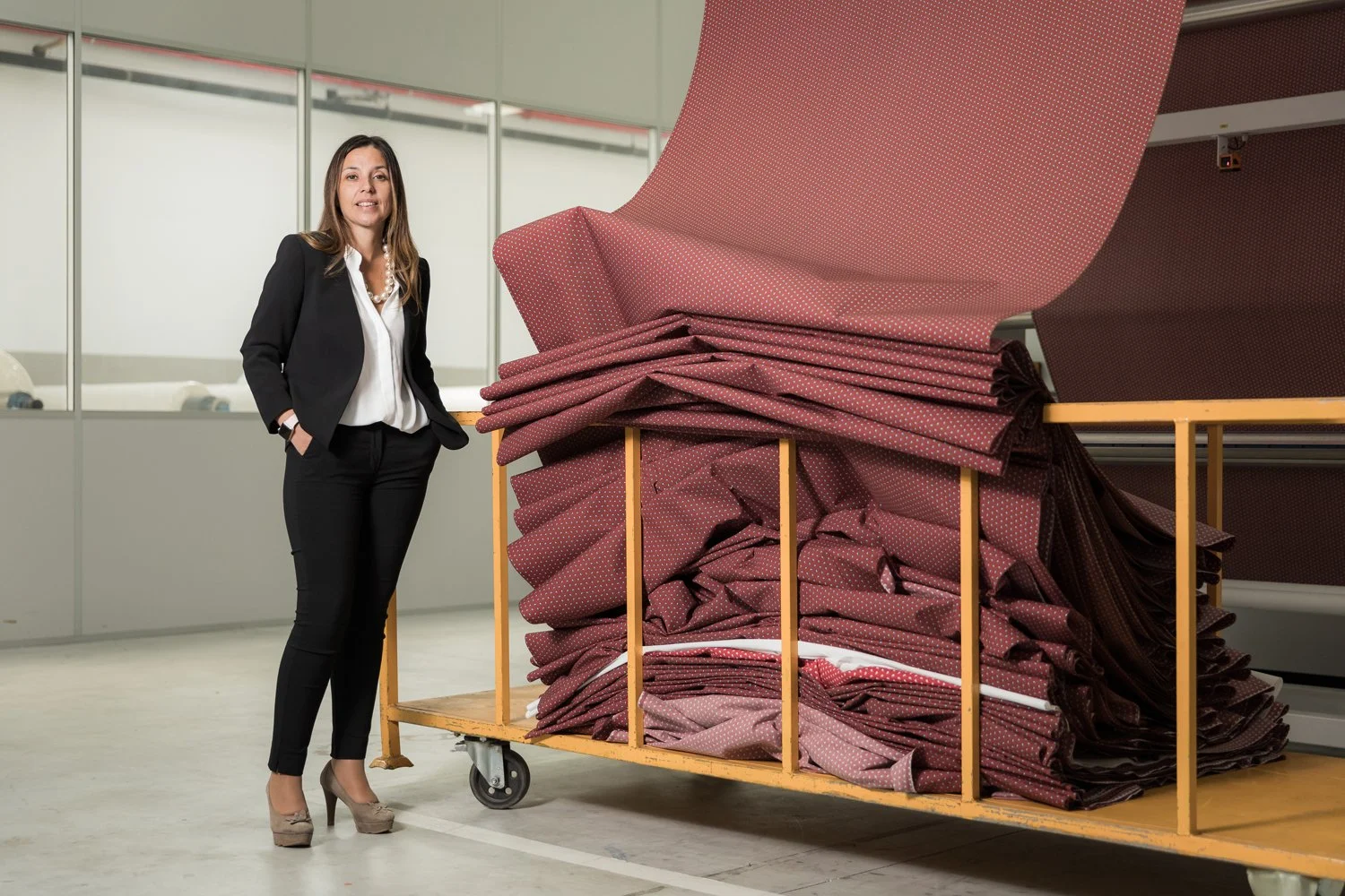 Mulher de negócios em traje preto e branco ao lado de uma pilha de tecidos vermelhos com pequenas pintas, em ambiente industrial.