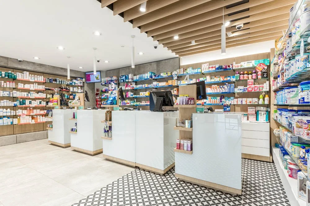 Interior de uma farmácia com balcões de atendimento, prateleiras com medicamentos e produtos de saúde, e telas de computador na área de caixa.