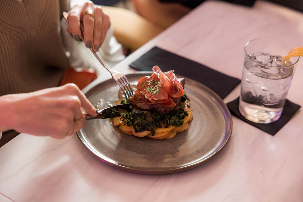 Pessoa segurando garfo e faca, servindo um prato de comida com carne, folhas verdes e uma porção de macarrão, em um restaurante.