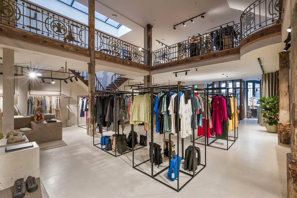 Interior de uma loja de roupas com estantes e cabides cheios de roupas coloridas, espaço bem iluminado e decorado com elementos de ferro envelhecido e plantas grandes.