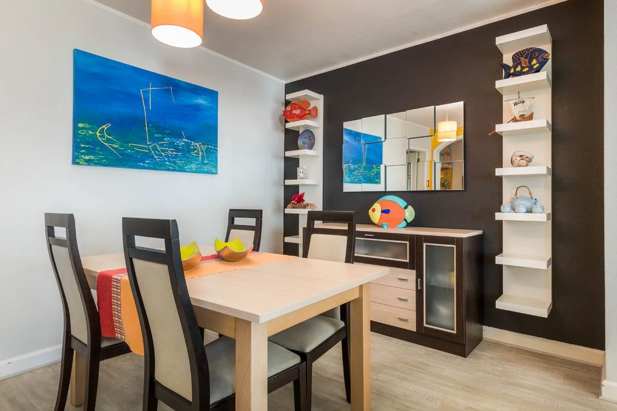 Sala de jantar decorada com paredes brancas e uma parede preta, mesa de jantar de madeira clara com cinco cadeiras pretas e almofadas brancas, espelho grande na parede preta, prateleiras brancas com enfeites, including peixes de pelúcia e vasos, quad