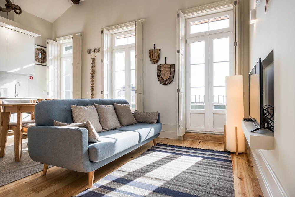 Sala de estar com sofá cinza, almofadas, televisão em parede branca, lampada de pé e janelas grandes com portas de vidro.