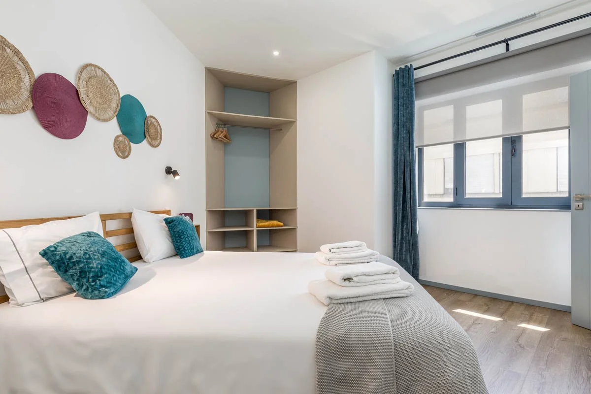 Quarto de hotel com cama de casal, roupa de cama branca, almofadas azuis, parede com decoração de cestos de fibra, closet com espaço para roupas e janela grande com cortinas azuis