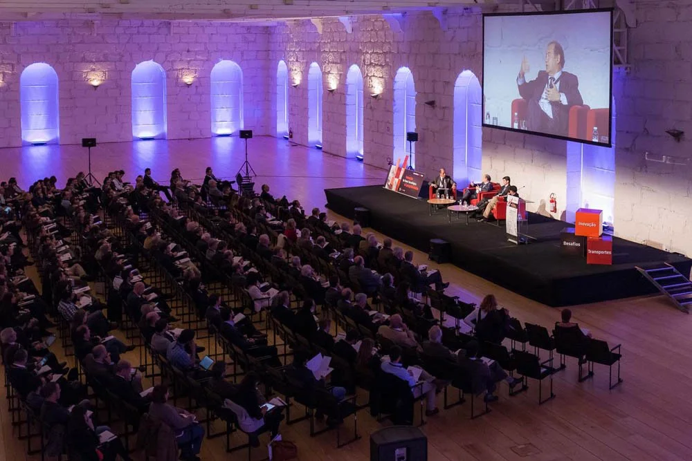 Conferência ou seminário com painel de palestrantes em palco, grande público sentado assistindo, cenário com paredes de pedra e iluminação azul, projeção de um palestrante na tela, o tema parece relacionado a inovação, desafios e transportes.