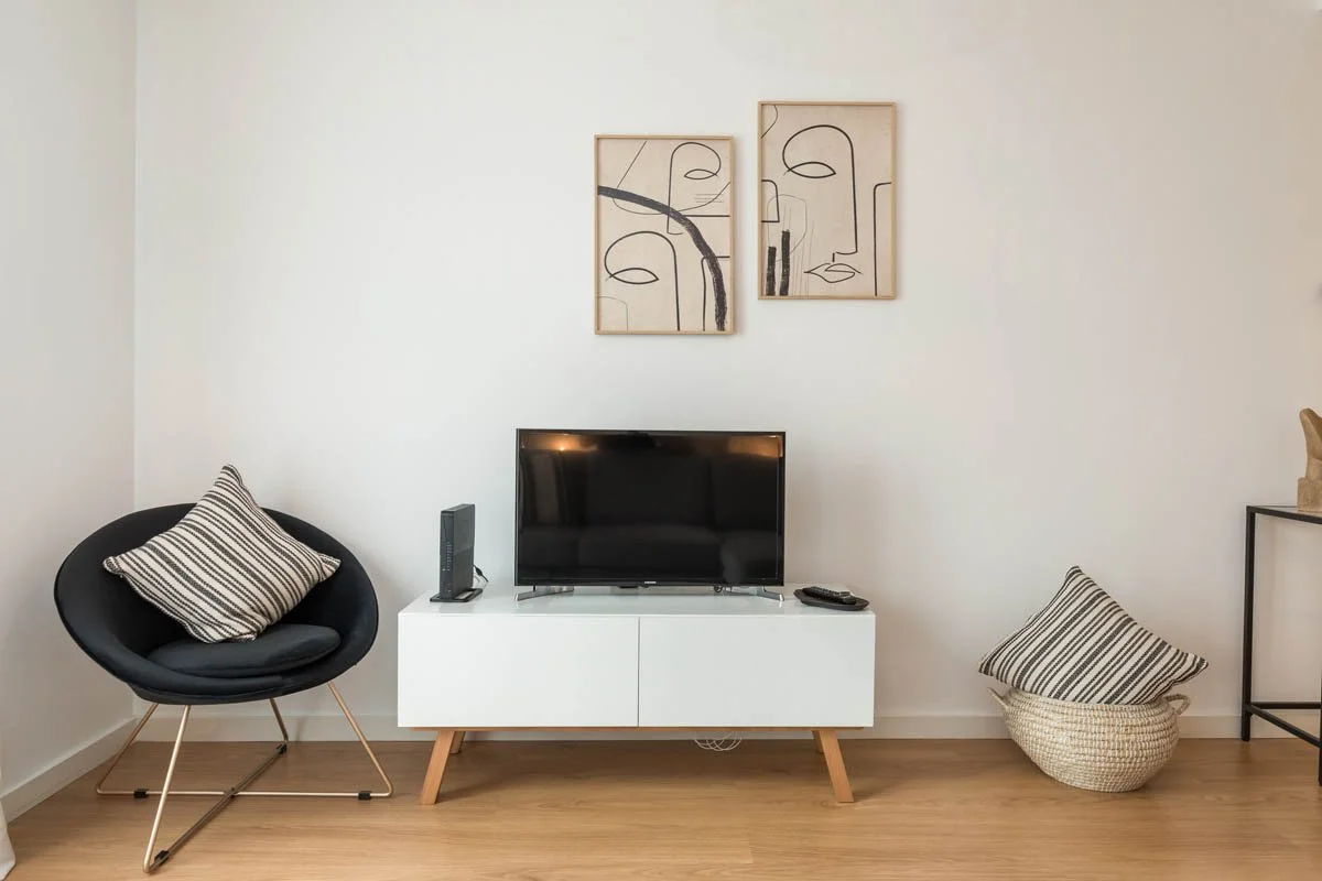 Sala de estar com televisão em uma mídia branca, duas almofadas com listras, uma em uma cadeira preta moderna e outra no cesto de fibra no chão, e uma parede branca com duas obras de arte minimalistas.