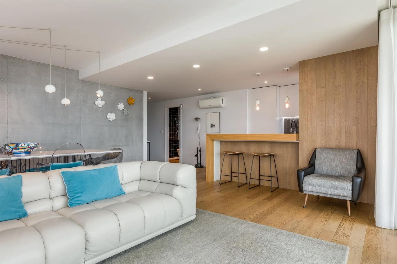 Sala de estar moderna com sofá branco, almofadas azuis, cadeira de descanso cinza e preto, balcão de bar com duas banquetas, parede de madeira, mesa de jantar com cadeiras, decorada com luzes pendentes e detalhes artísticos na parede