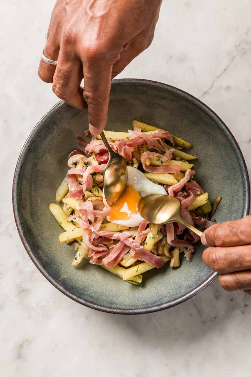 Prato com batatas, presunto, cogumelos, e ovo poché sendo temperado com colheras em uma tigela cinza escura