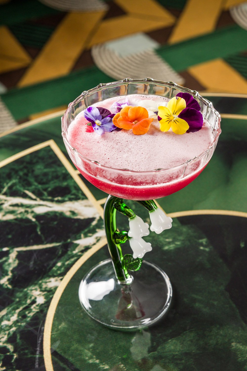 Coquetel rosa em taça decorada com flores comestíveis roxas, amarelas e laranja, em uma mesa de mármore verde escuro.