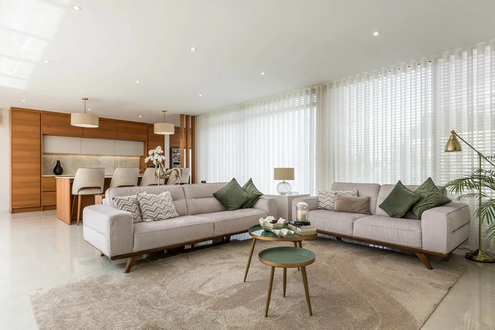 Sala de estar com dois sofás cor de creme, uma mesa de centro com objetos decorativos, cortinas brancas e iluminação suave.