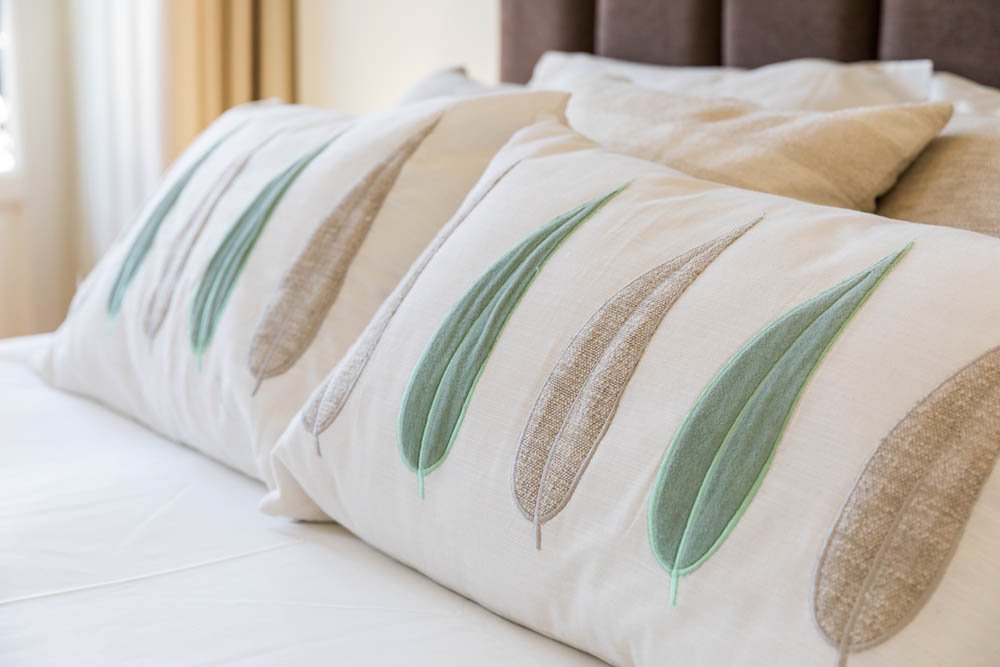 Três travesseiros com capa branca e estampa de penas verdes e marrons na cama, com cabeceira de madeira ao fundo.