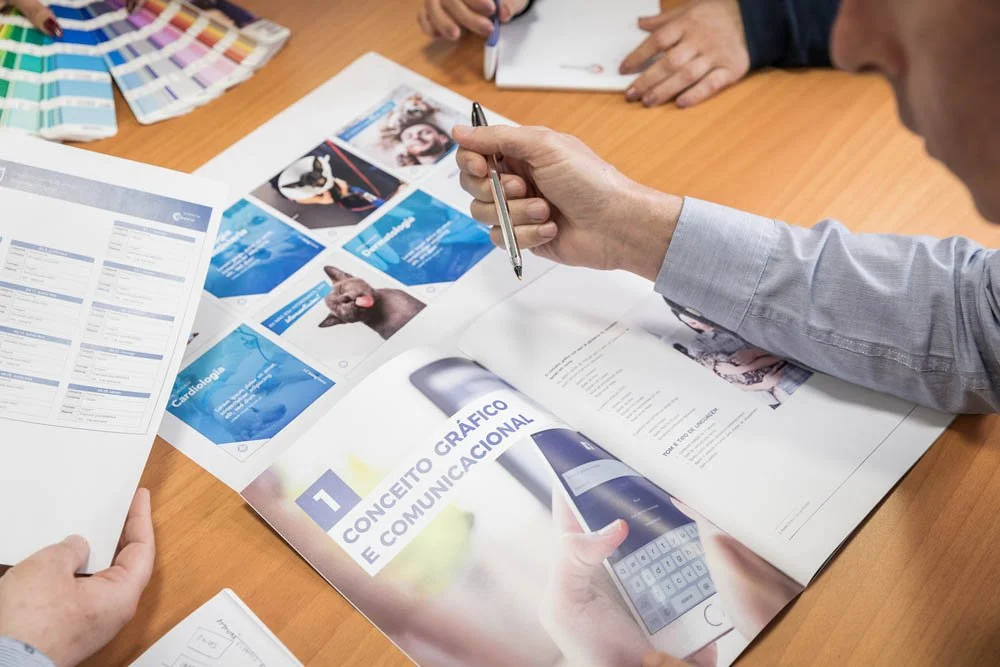 Homem em uma reunião de negócios ou projeto, analisando materiais impressos e digitando em um laptop, envolvendo comunicação visual e estratégias de marketing.