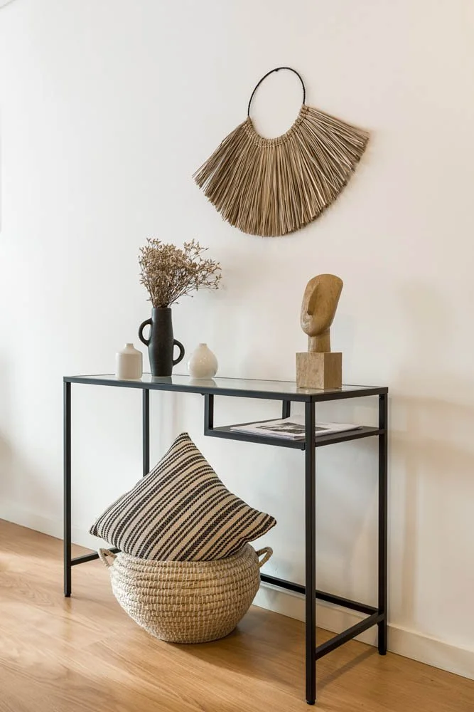 Mesa de metal preta com decoração minimalista: vasos, busto de madeira, e revista. Cesto de vime com travesseiro geométrico. Quadro na parede e decoração de palha rústica.