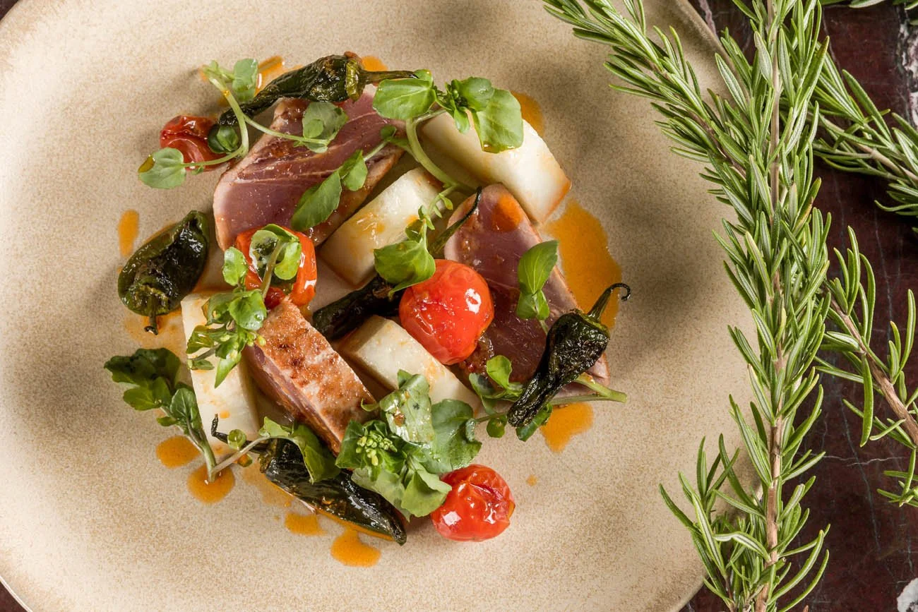 Prato de carne com vegetais, tomate, folhas verdes e molho servido em prato de cerâmica, ao lado ramos de alecrim.