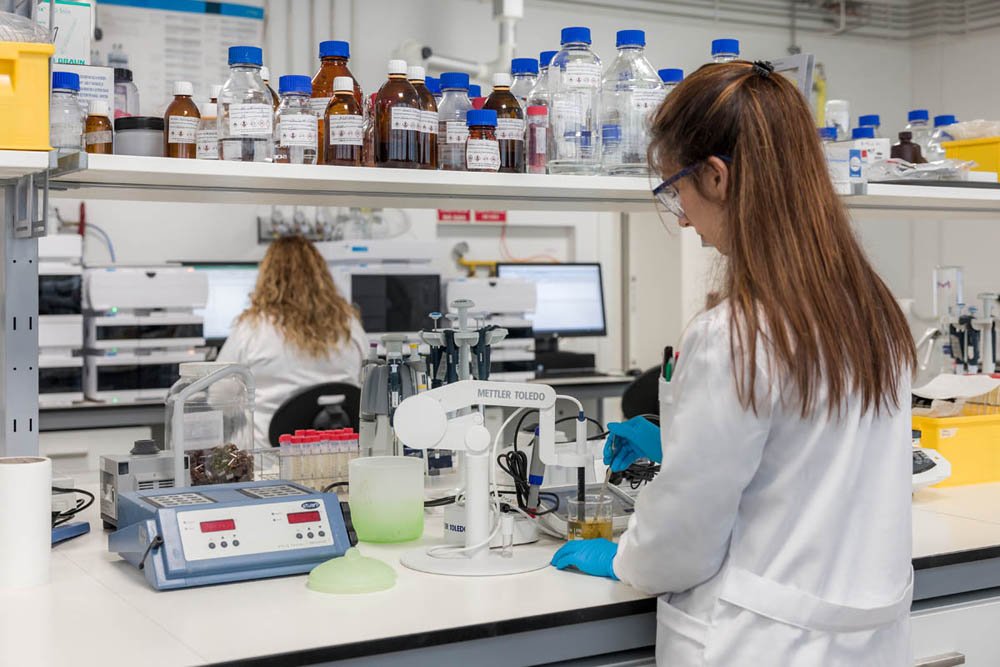 Pesquisadora usando jaleco e óculos de proteção em um laboratório, manipulando um recipiente com líquidos, com diversos vidrarias, equipamentos de laboratório e computadores ao fundo.