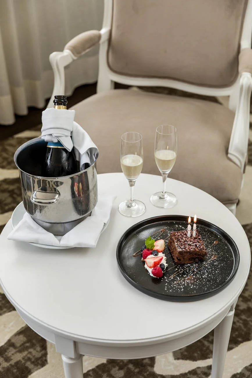 Mesa com uma garrafa de champanhe, duas taças de espumante, e uma sobremesa de bolo de chocolate com velas de aniversário, decorada com frutas vermelhas, folhas de hortelã, açúcar de confeiteiro, e um sabonete de chocolate.