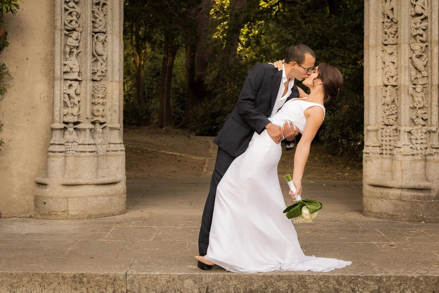 Casal de casamento em um abraço romântico diante de uma estrutura de pedra com detalhes decorativos, com árvores ao fundo.