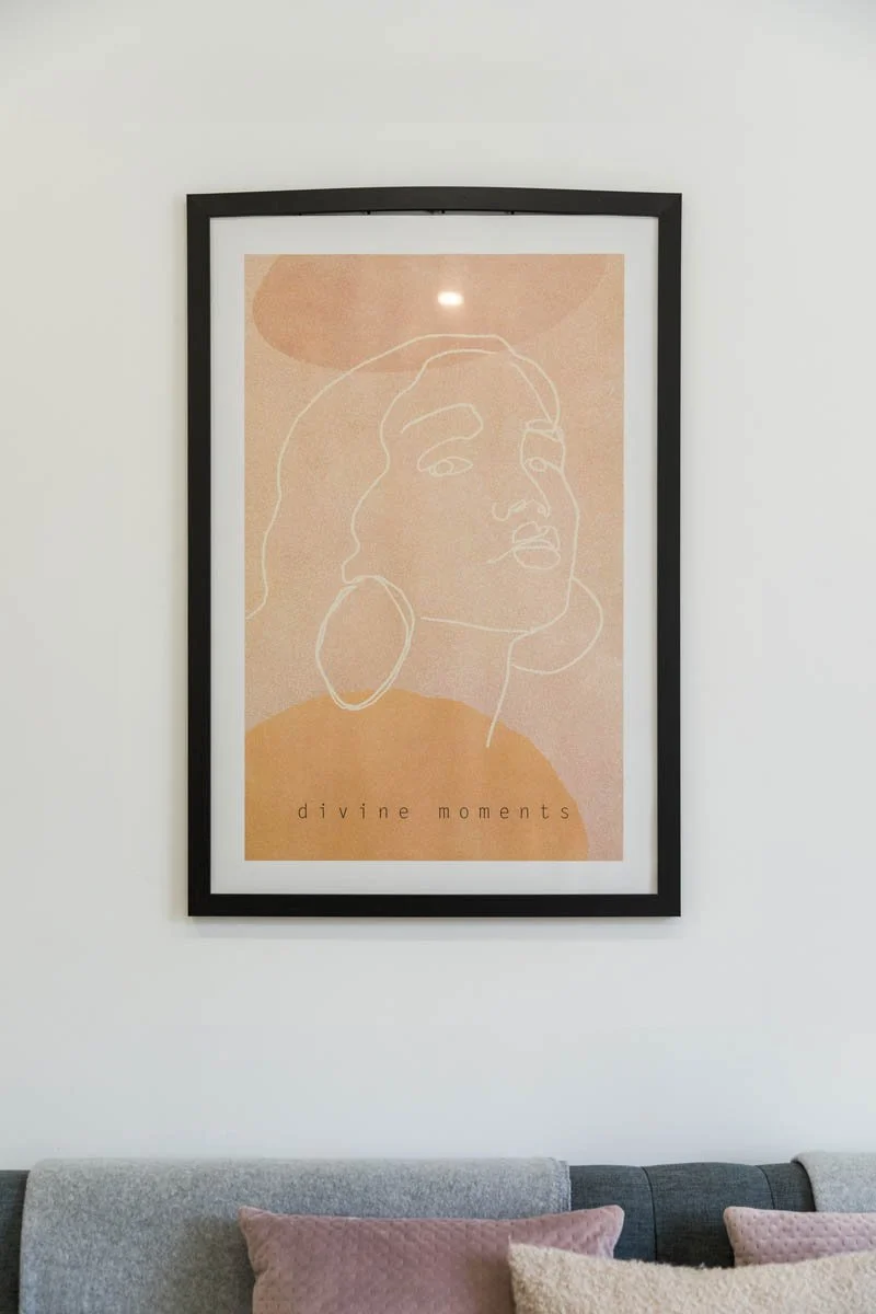 Quadro decorativo com arte de linha de uma mulher e a frase "divine moments" na parede de uma sala com sofá e almofadas.