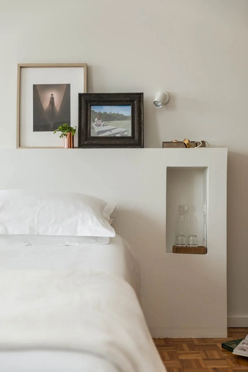 Cabeceira de cama branca com quadros decorativos na parede, uma pequena planta em um vaso cobre rose e uma prateleira embutida com dois copos ao lado de uma garrafa na parede, piso de madeira.