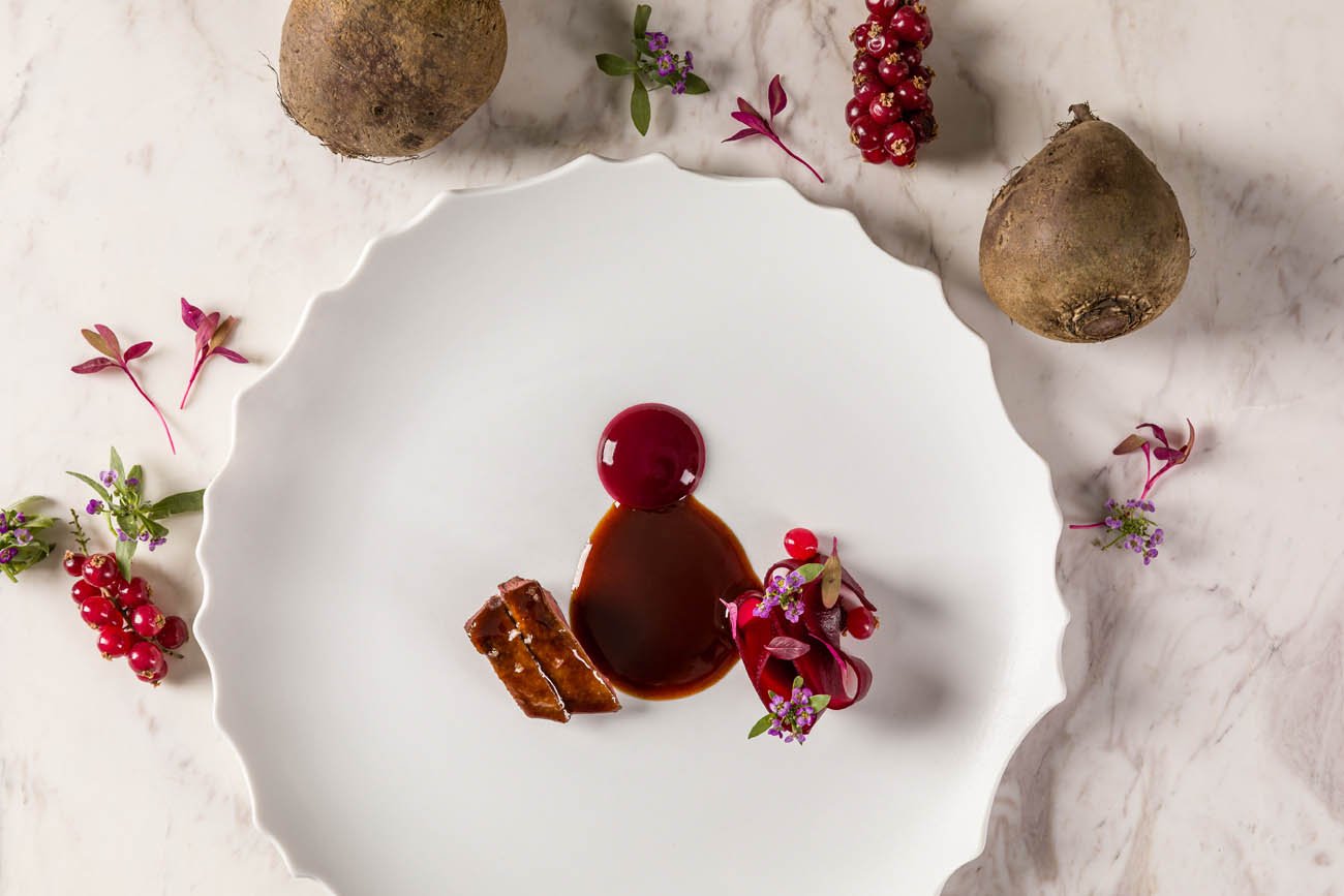 Prato branco decorado com molho, uma cereja, pedaço de doce e flores pequenas, ao redor de batatas e frutas vermelhas.