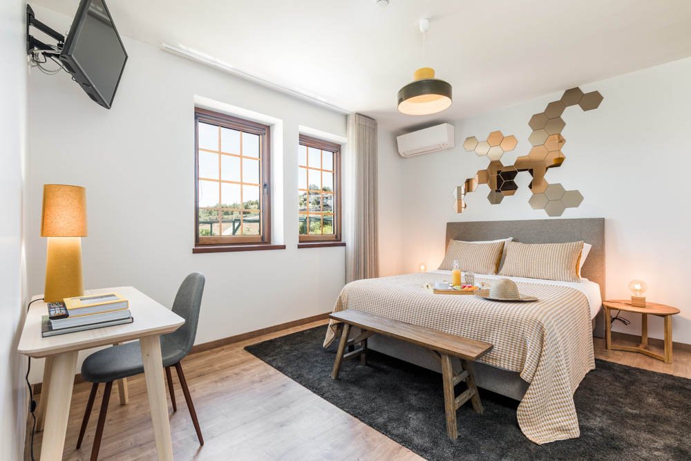 Quarto de hotel com cama, parede decorada com espelhos hexagonais, janela com cortina, televisão na parede, luminária de chão ao lado da mesa, janela com vista para o exterior, parede com ar-condicionado, e iluminação suave.